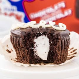 A chocolate cupcake with cream filling and white icing on top sits in a wrapper, with a large bite taken out, revealing the creamy center—just like classic Hostess Cupcakes.