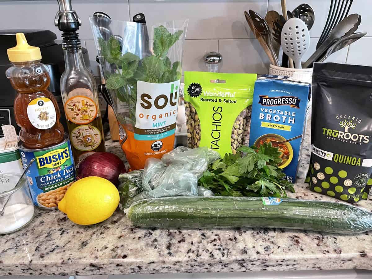 Various ingredients on a kitchen counter, including honey, olive oil, mint, pistachios, chickpeas, vegetable broth, quinoa, a lemon, cucumber, an onion, parsley, and a bag of greens—perfect for preparing the fresh and vibrant Jennifer Aniston Salad.