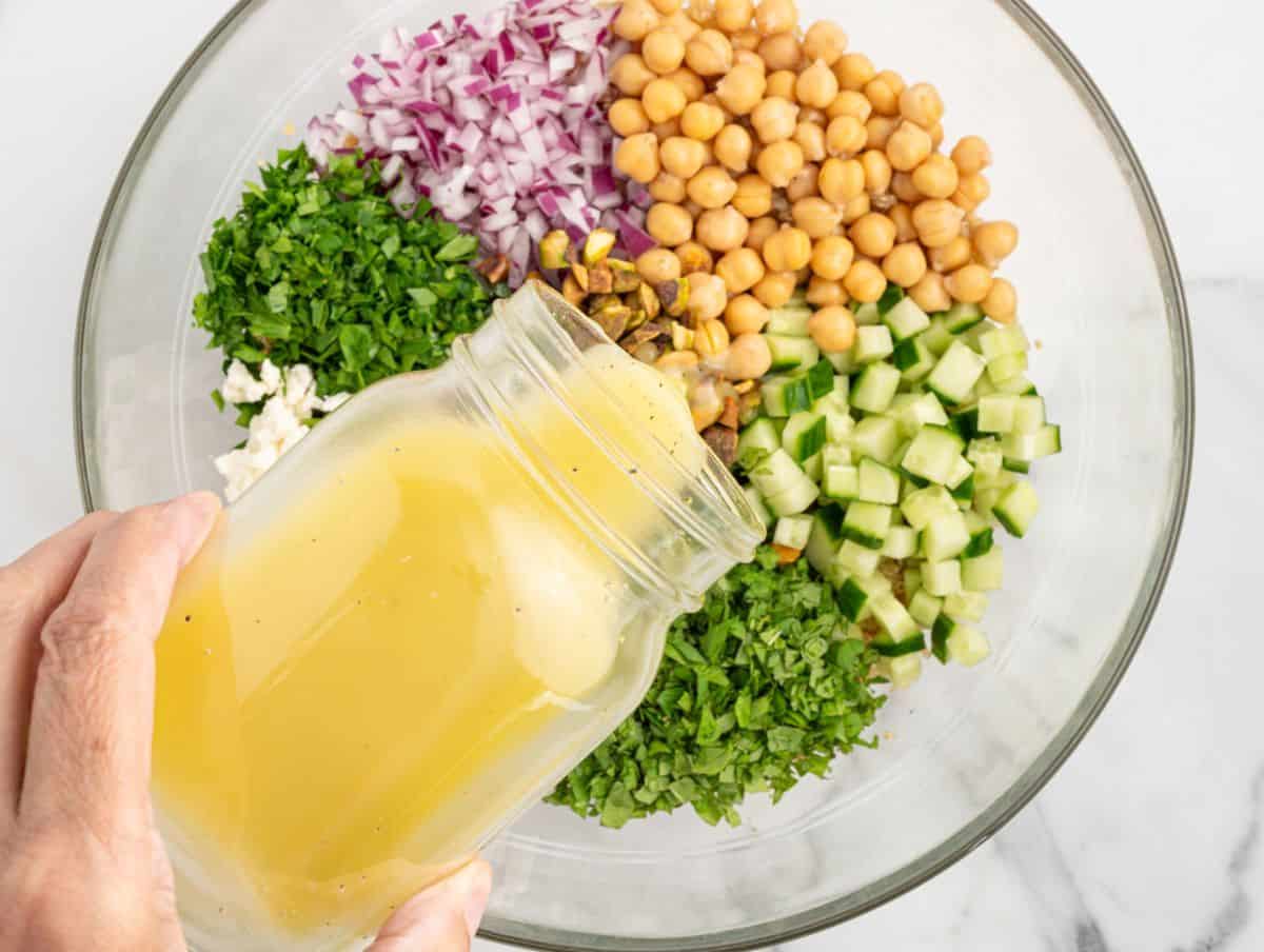 A hand pours yellow dressing from a jar into a bowl with chickpeas, chopped cucumber, red onion, herbs, feta, and pistachios—classic ingredients for a vibrant Jennifer Aniston Salad—ready to be mixed for a wholesome salad.