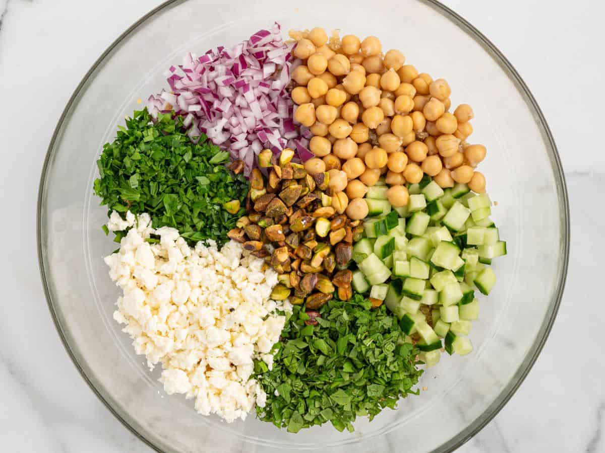 A glass bowl with sections of chopped red onion, chickpeas, cucumber, parsley, mint, feta cheese, and pistachios on a white surface—classic ingredients that make up the vibrant Jennifer Aniston Salad.