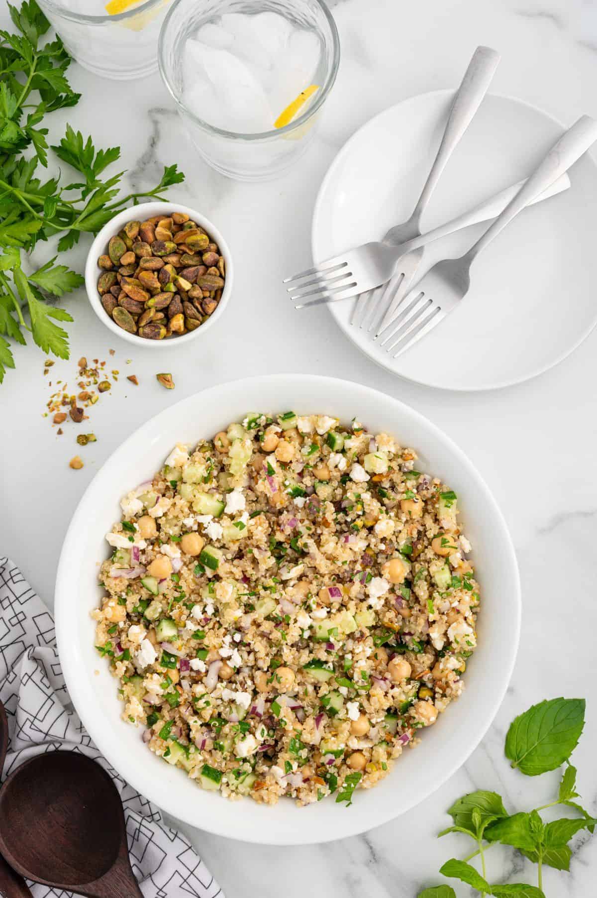 A bowl of quinoa salad inspired by the Jennifer Aniston Salad, featuring chickpeas, feta, herbs, and red onion, sits on a white table surrounded by plates, forks, a small bowl of pistachios, and glasses of water with lemon slices.