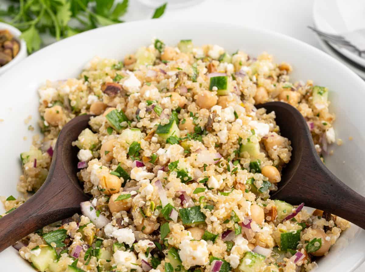A bowl of quinoa salad with chickpeas, cucumbers, red onions, feta cheese, and herbs—reminiscent of the famous Jennifer Aniston Salad—is being served with wooden salad servers.
