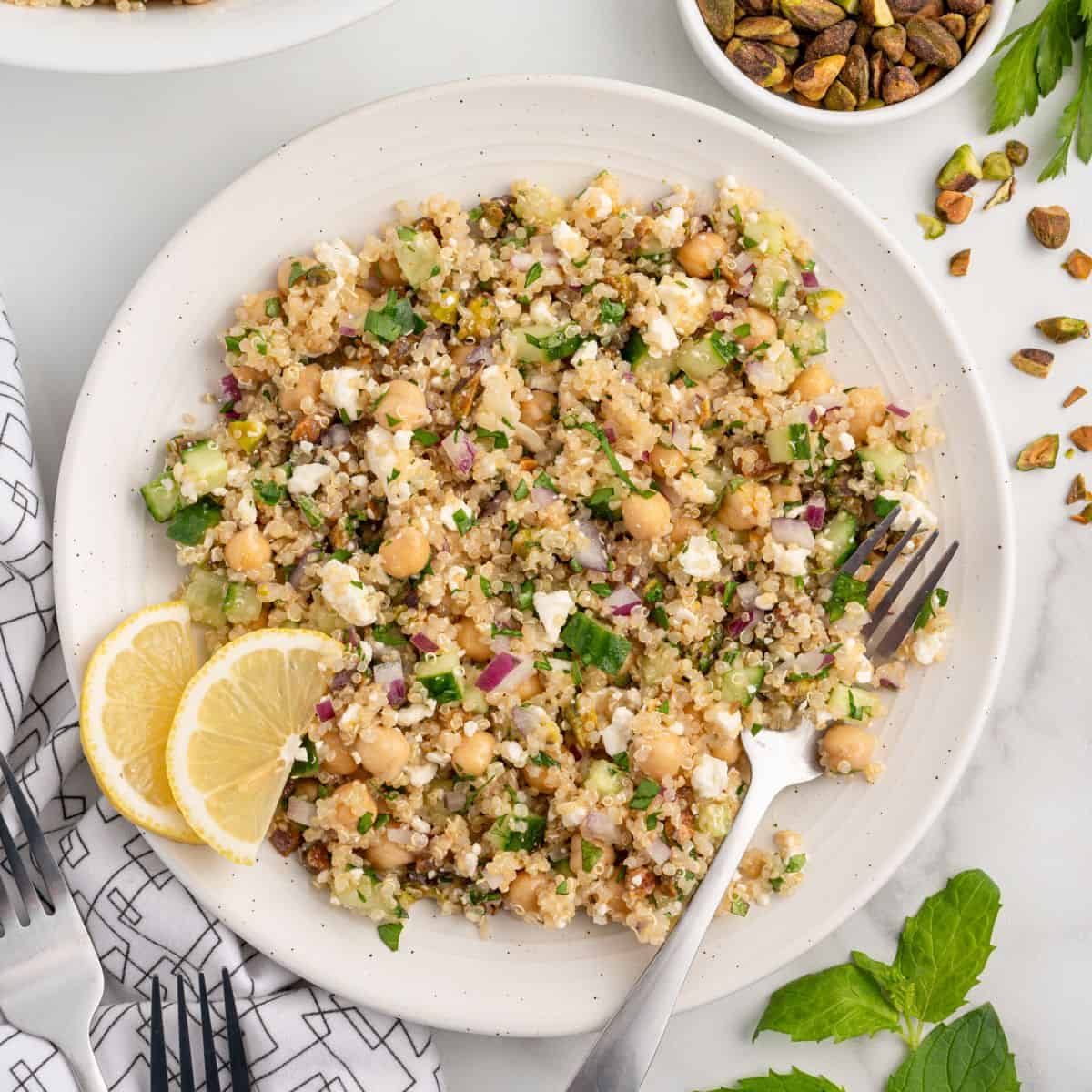 A plate of quinoa salad with chickpeas, feta, red onion, cucumber, herbs, and lemon slices—often known as the Jennifer Aniston Salad—is served with a fork on the plate. A bowl of pistachios sits in the corner.