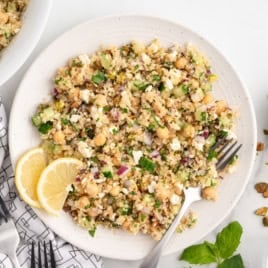 A plate of quinoa salad with chickpeas, chopped vegetables, feta cheese, and herbs—often reminiscent of the famed Jennifer Aniston Salad—is garnished with two lemon slices. A fork rests on the plate; pistachios and fresh greens are nearby.