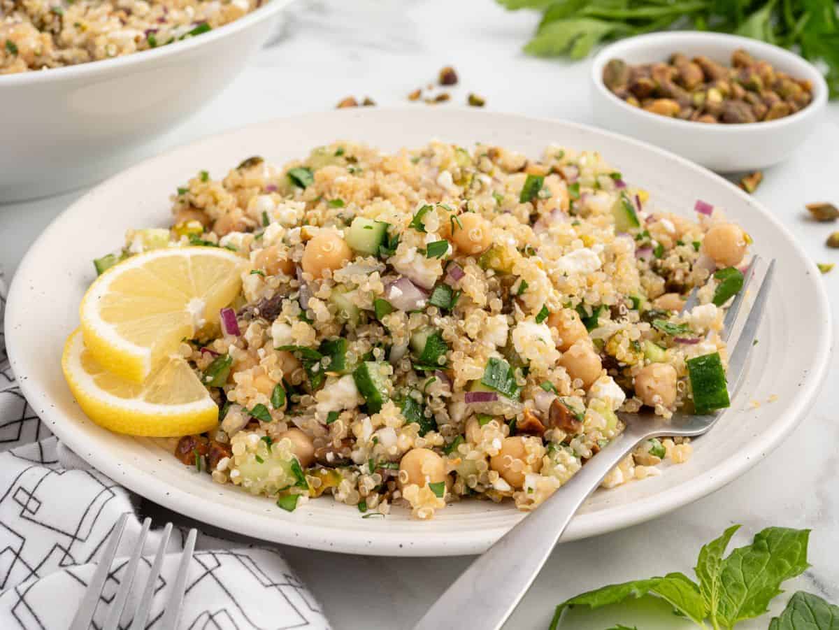 A plate of quinoa salad with cucumber, chickpeas, red onion, and herbs, garnished with lemon slices—reminiscent of the popular Jennifer Aniston Salad. A fork rests on the plate, while fresh greens and pistachios are visible in the background.