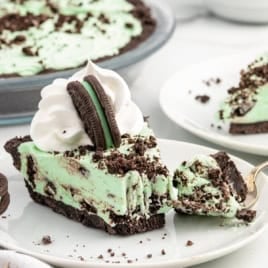 A slice of Mint Chocolate Pie with whipped cream and a chocolate sandwich cookie on top, served on a white plate, with cookie crumbs scattered around.