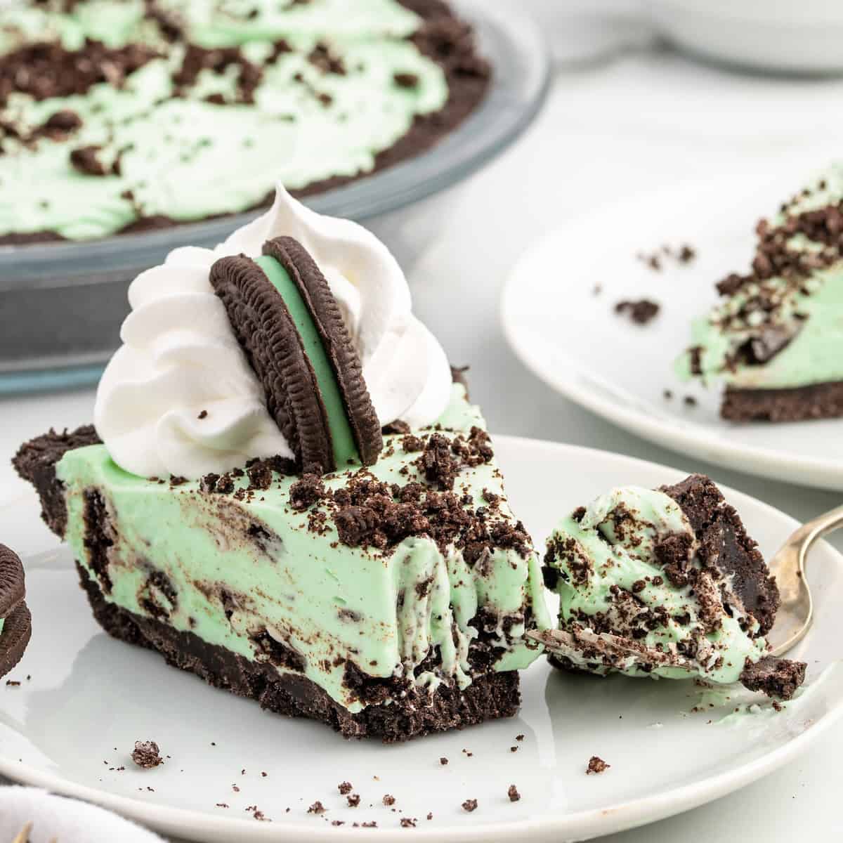 A slice of Mint Chocolate Pie with whipped cream and a chocolate sandwich cookie on top, served on a white plate, with cookie crumbs scattered around.