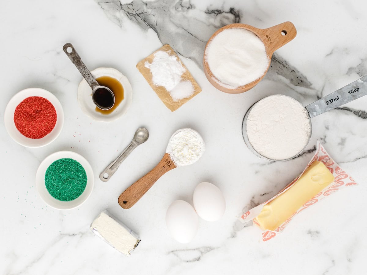 Baking ingredients on a marble surface: flour, sugar, butter, eggs, baking soda, vanilla, cream cheese, red and green sprinkles—just what you need for Grandma’s Christmas Sugar Cookies Recipe—all perfectly measured in bowls and spoons.