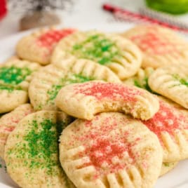 A plate of Christmas Sugar Cookies topped with red and green sprinkles. One cookie has a bite taken out of it, showing a soft interior. Other cookies are stacked around it, just like Grandma’s Christmas Sugar Cookies Recipe always delivers—festive and irresistibly soft.