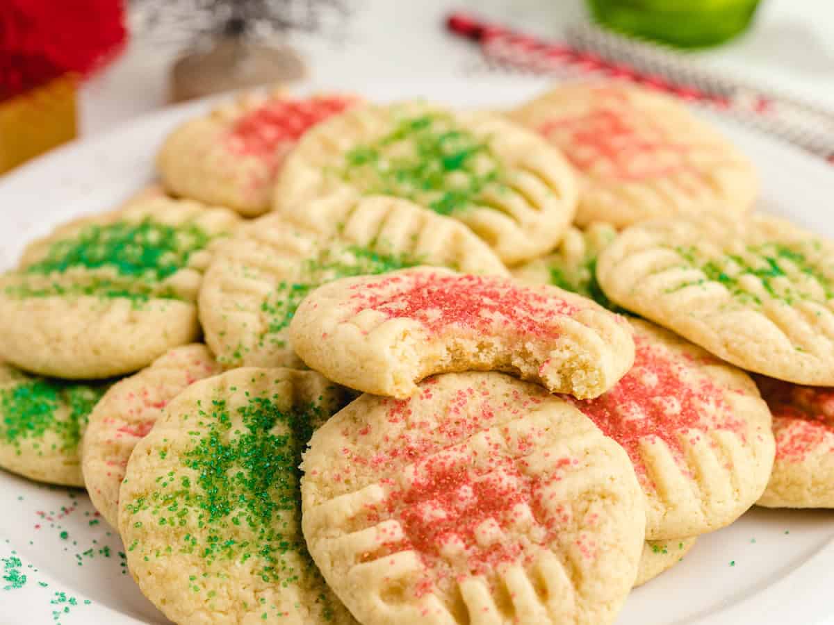 A plate of Christmas Sugar Cookies topped with red and green sprinkles. One cookie has a bite taken out of it, showing a soft interior. Other cookies are stacked around it, just like Grandma’s Christmas Sugar Cookies Recipe always delivers—festive and irresistibly soft.