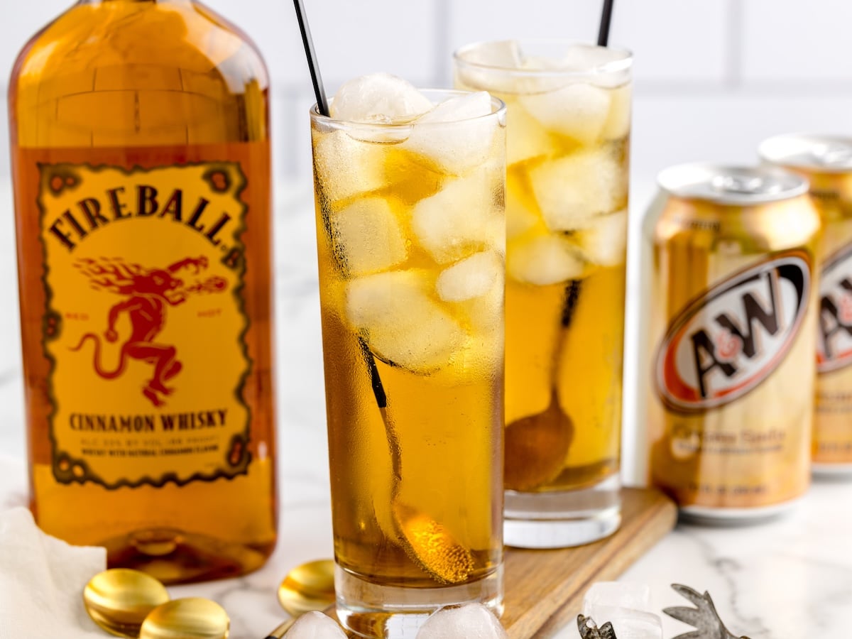 Two tall glasses filled with ice and a golden-brown Fireball and Cream Soda sit beside a bottle of Fireball Cinnamon Whisky and two cans of A&W Root Beer on a white surface.