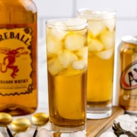 Two tall glasses filled with ice and an amber-colored Fireball and Cream Soda drink sit next to a Fireball Cinnamon Whisky bottle and two A&W cream soda cans on a wooden board. Spoons and ice cubes are visible in the foreground.