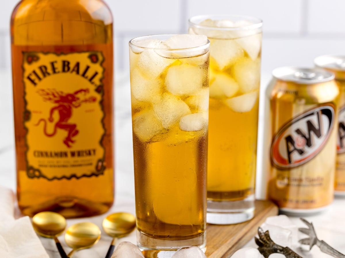 Two tall glasses filled with ice and an amber-colored Fireball and Cream Soda drink sit next to a Fireball Cinnamon Whisky bottle and two A&W cream soda cans on a wooden board. Spoons and ice cubes are visible in the foreground.