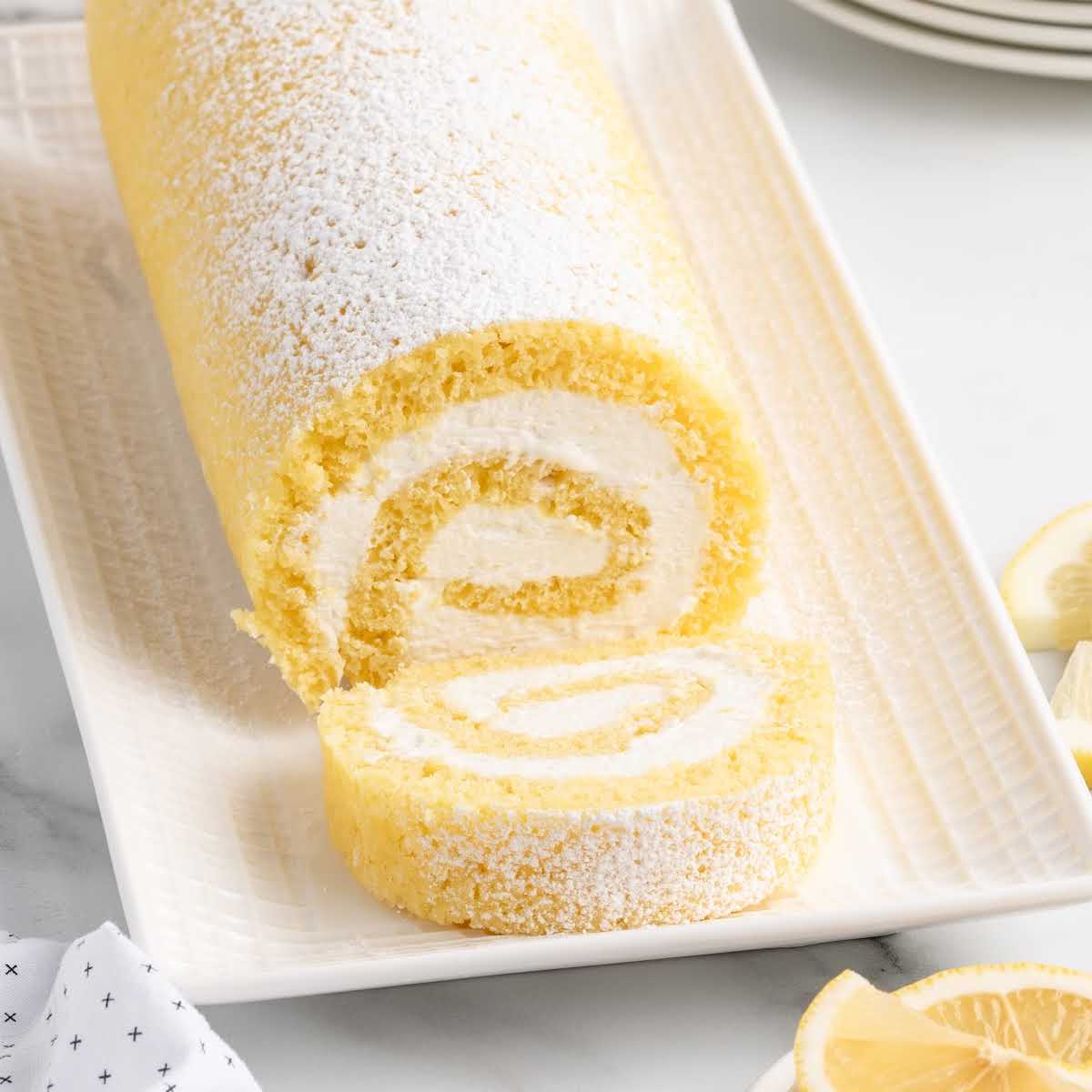 Lemon swiss roll cake served on a rectangular plate with lemon wedges and dessert forks nearby