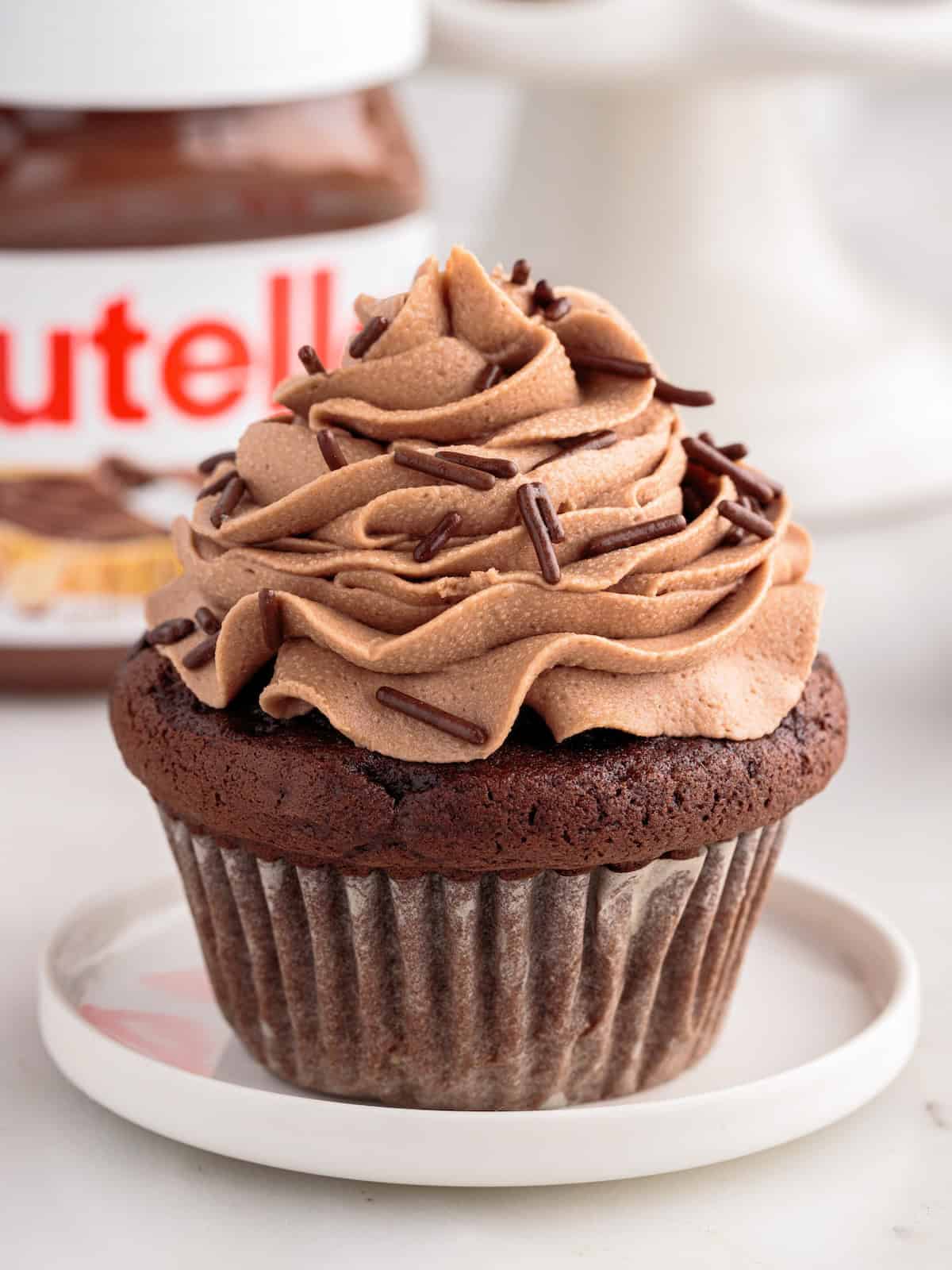 A decadent Nutella Cupcake with swirled chocolate frosting and chocolate sprinkles sits on a small plate. A jar of Nutella is blurred in the background, hinting at the rich, hazelnut flavor baked into every bite.
