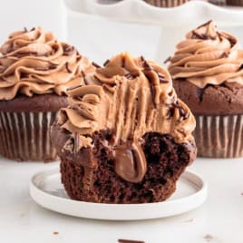 A Nutella Cupcake with chocolate frosting and filling is missing a bite, sitting on a plate. Two whole, similarly decorated Nutella Cupcakes are in the background.
