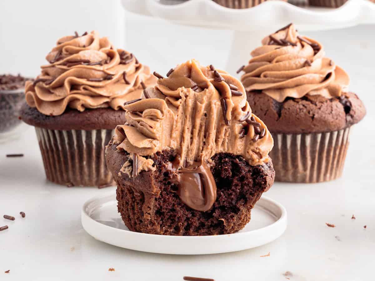 A Nutella Cupcake with chocolate frosting and filling is missing a bite, sitting on a plate. Two whole, similarly decorated Nutella Cupcakes are in the background.