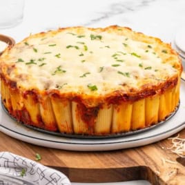 A delightful Honeycomb Pasta Bake, featuring upright rigatoni noodles topped with melted cheese and herbs, sits on a white plate placed on a wooden board, surrounded by grated cheese and a bowl of sauce.