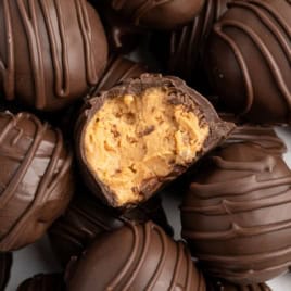 A close-up of chocolate truffles, with one cut open to reveal a creamy peanut butter filling reminiscent of Peanut Butter Balls with Rice Krispies. The truffles are drizzled with chocolate and stacked together.