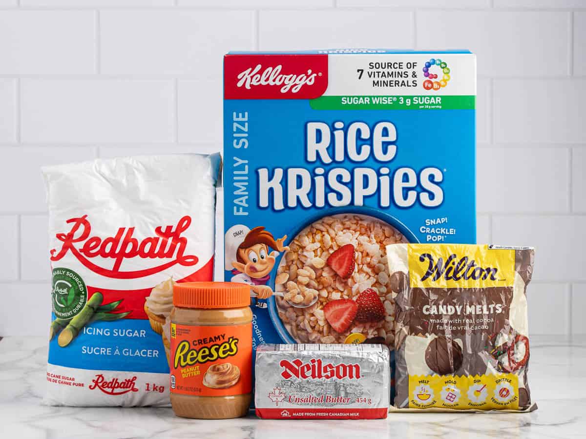 A bag of Redpath icing sugar, a jar of Reeseโs peanut butter, a box of Kelloggโs Rice Krispies, a block of Neilson butter, and a bag of Wilton chocolate candy melts are displayed on a white counterโeverything you need to make delicious Peanut Butter Balls with Rice Krispies.
