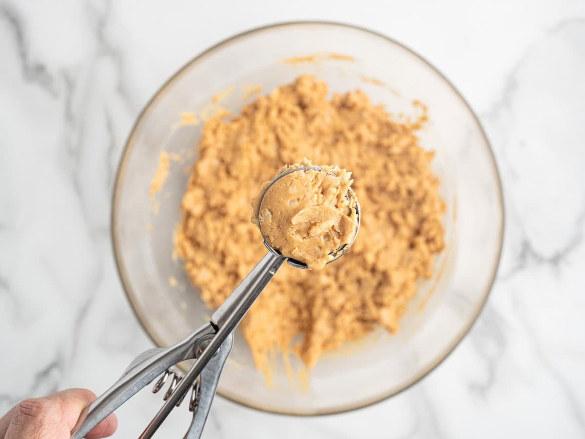 A hand holds a cookie scoop with raw dough for Peanut Butter Balls with Rice Krispies above a glass bowl filled with more dough, all on a white marble surface.