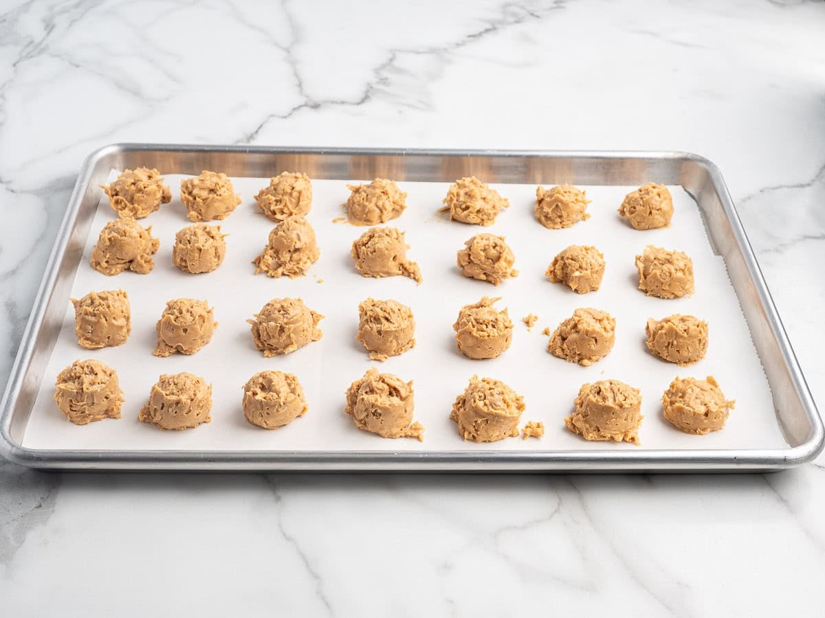 A baking sheet lined with parchment paper holds 24 evenly spaced scoops of raw cookie dough, reminiscent of Peanut Butter Balls with Rice Krispies, ready to be baked. The background is a white marble countertop.