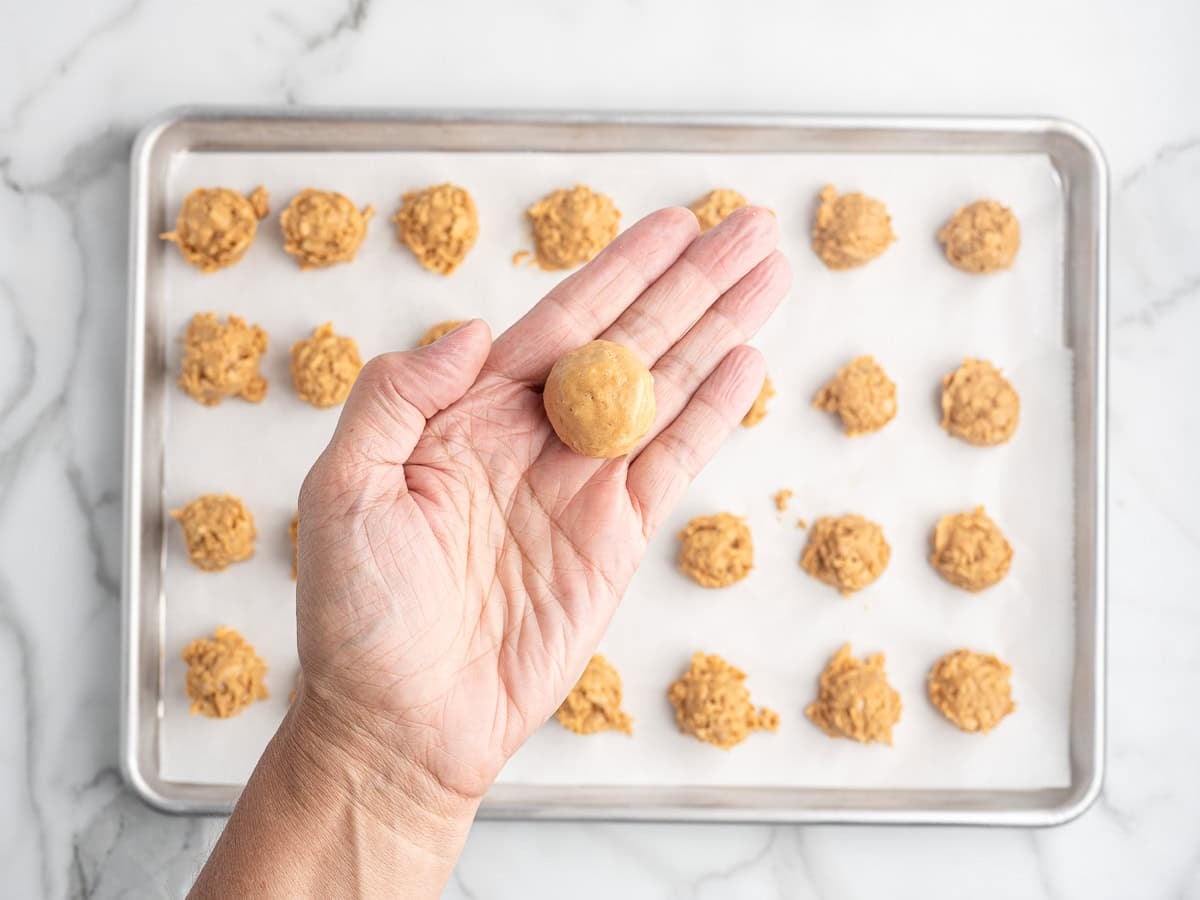 A hand holds a round ball of Peanut Butter Balls with Rice Krispies cookie dough above a baking sheet lined with parchment paper, with more dough portions arranged on the tray in rows.