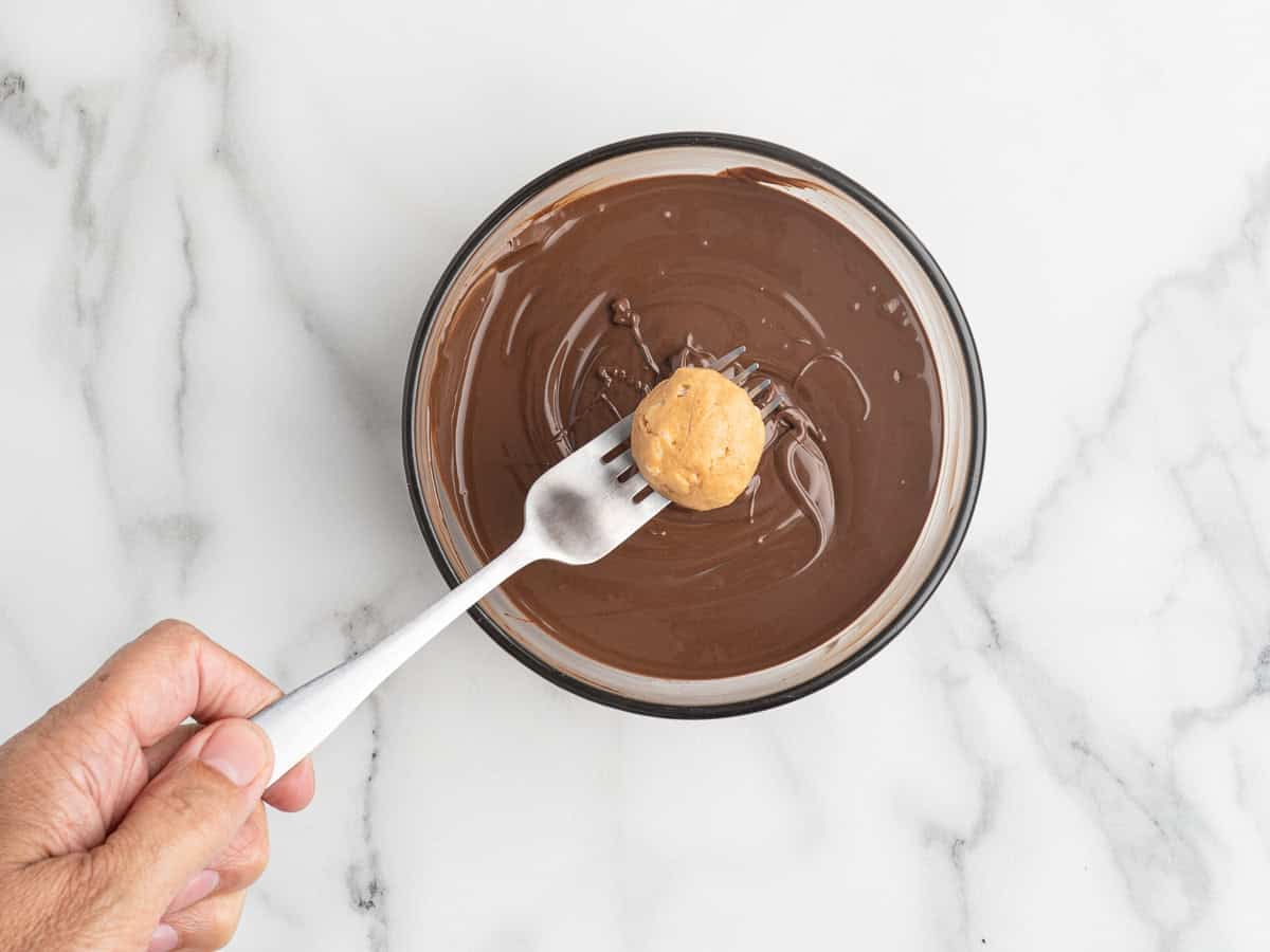 A hand holds a fork with a Peanut Butter Balls with Rice Krispies mixture over a bowl of melted chocolate on a white marble surface.