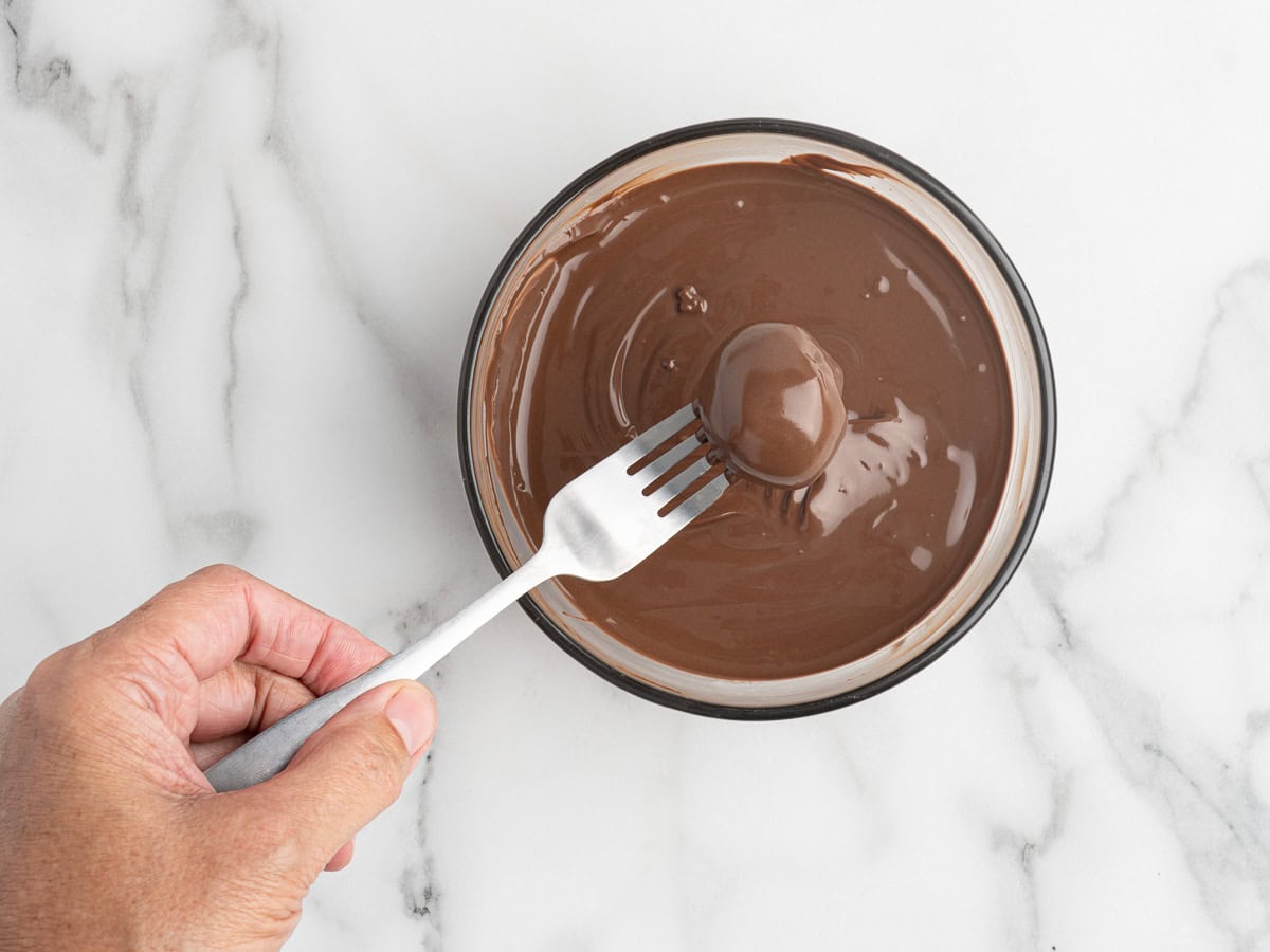 A hand holding a fork dips a round Peanut Butter Ball with Rice Krispies into a bowl of melted chocolate on a white marble surface.