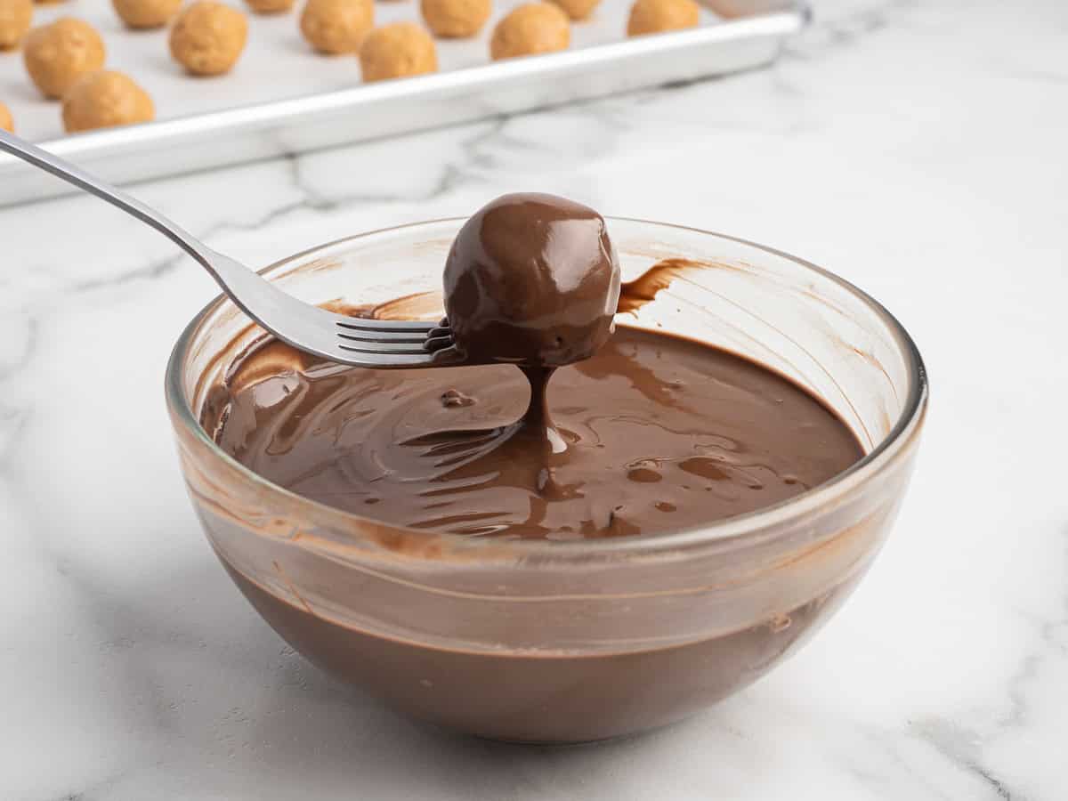 A fork holds a round Peanut Butter Ball with Rice Krispies dipped in melted chocolate above a glass bowl, with more undipped treats resting on a baking sheet in the background.