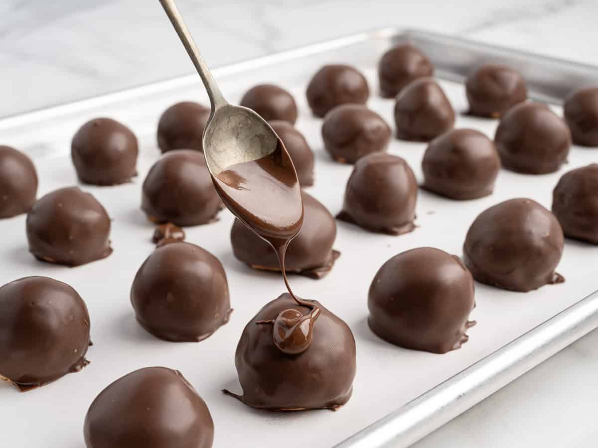 A spoon drizzles chocolate over a round Peanut Butter Ball with Rice Krispies on a baking sheet lined with parchment paper, with several other treats in the background.