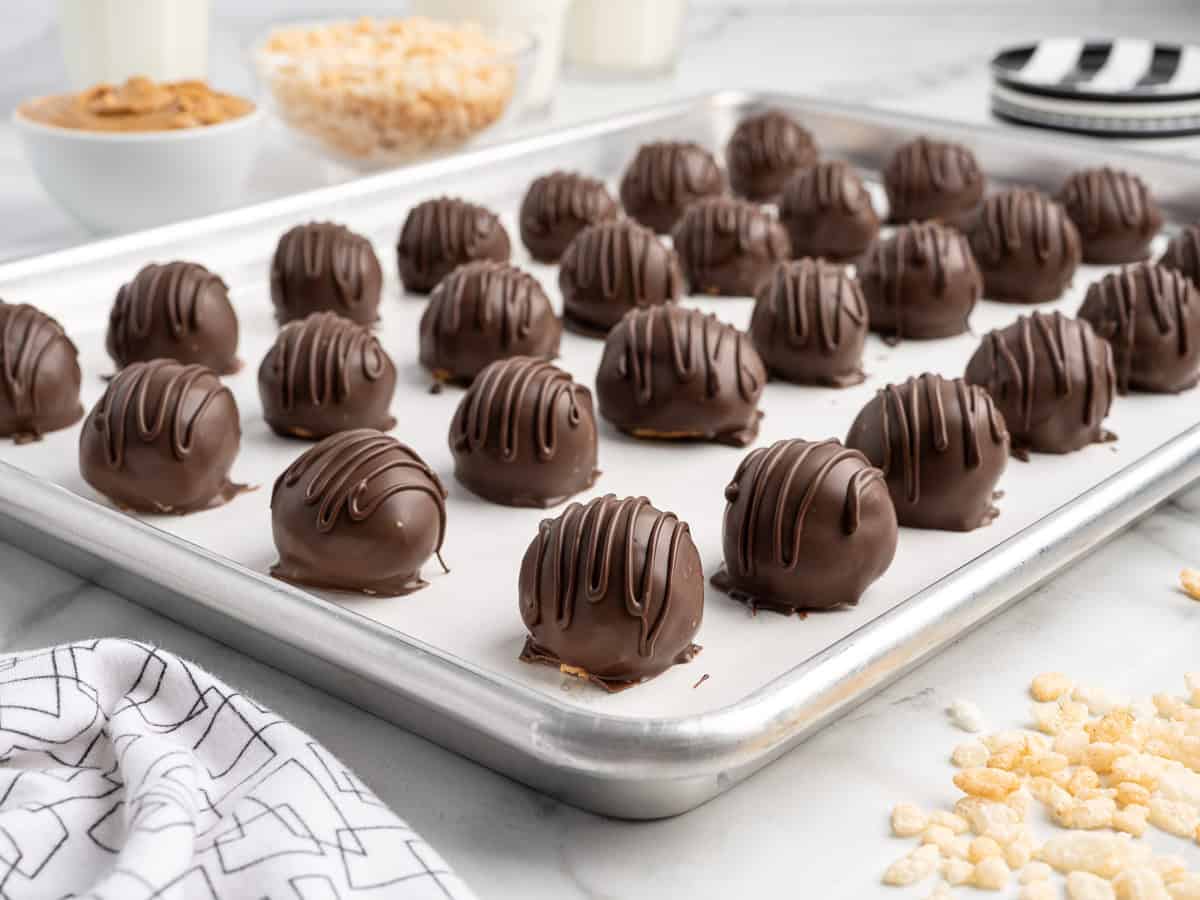 A baking tray with rows of round chocolate truffles drizzled with chocolate, set on parchment paper. In the background are bowls of cereal and peanut butter, and a glass of milkโperfect ingredients for making Peanut Butter Balls with Rice Krispies.