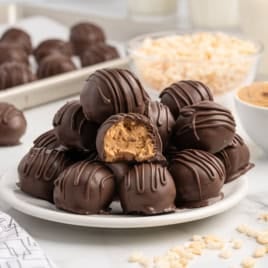A plate of chocolate-coated Peanut Butter Balls with Rice Krispies, one with a bite taken out, sits on a table with bowls of peanut butter, puffed rice, and more treats in the background.