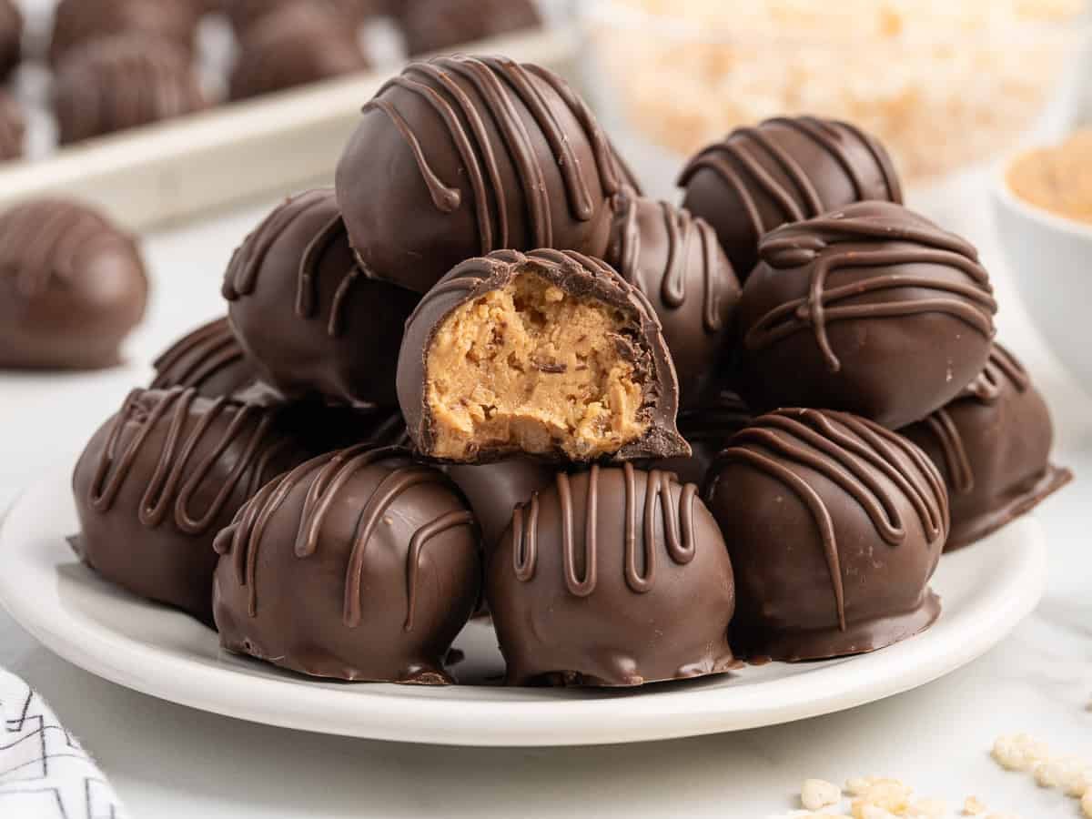 A plate of chocolate truffles with a drizzle on top is shown. One truffle is cut open, revealing a creamy, light brown peanut butter filling insideโsimilar to the irresistible Peanut Butter Balls with Rice Krispies. More truffles and ingredients are blurred in the background.
