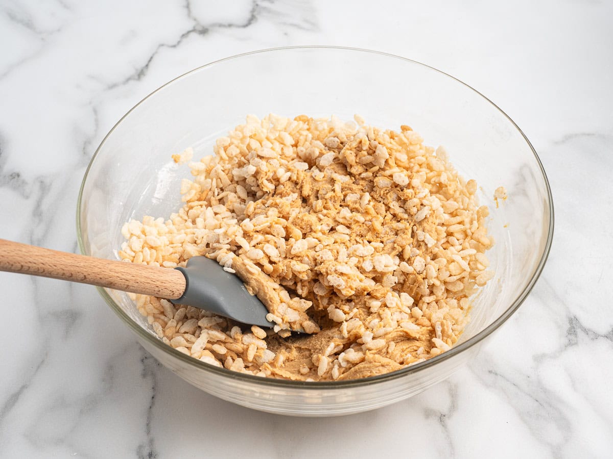 A glass bowl with rice cereal and a sticky mixture for Peanut Butter Balls with Rice Krispies is being stirred with a gray spatula, sitting on a white marble countertop.
