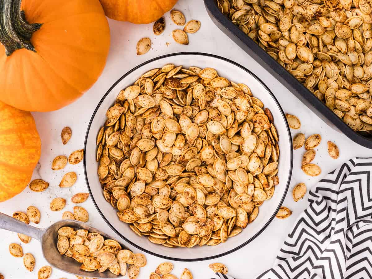 A bowl of seasoned Air Fryer Pumpkin Seeds sits on a table with whole pumpkins, a scoop of seeds, a baking tray of seeds, and a zigzag-patterned cloth nearby.