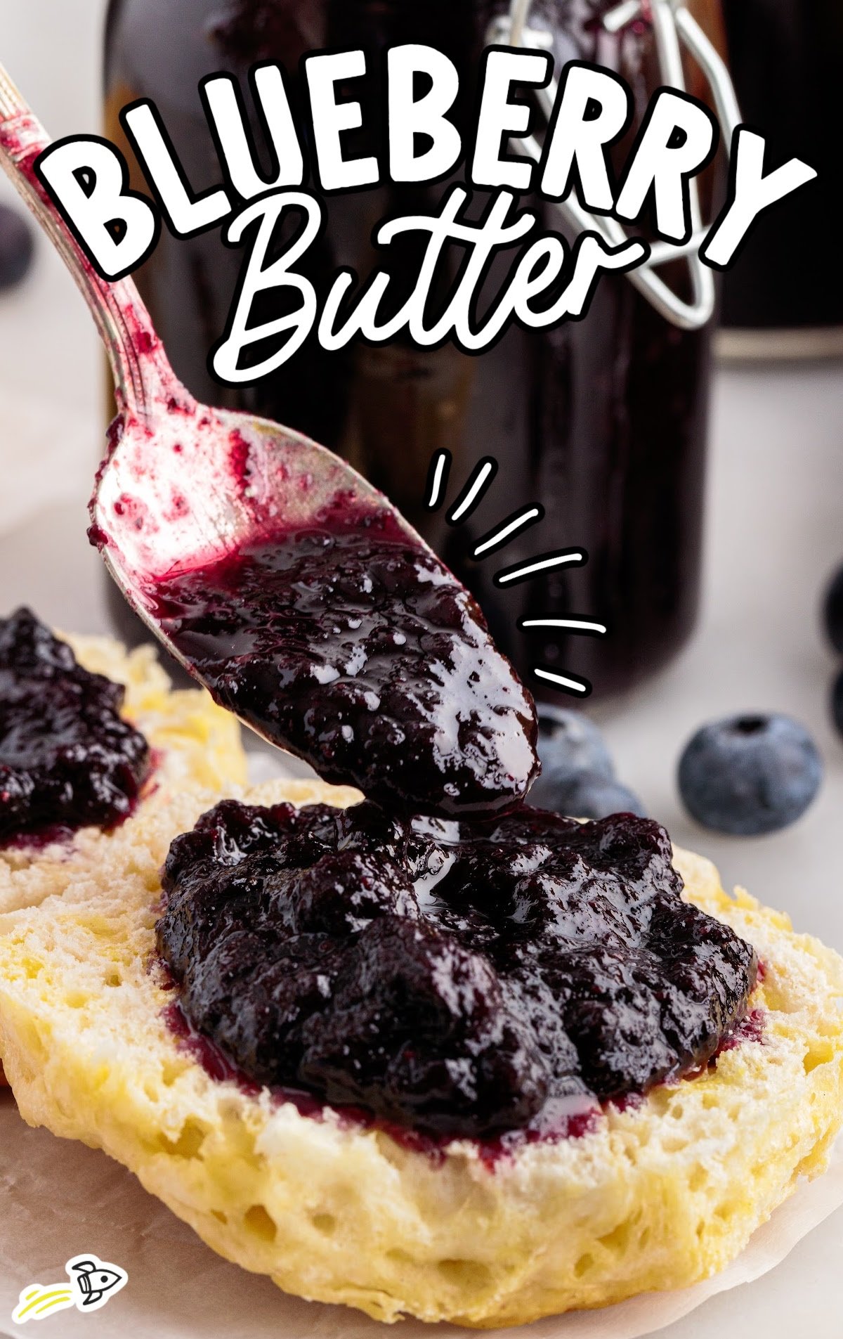 Homemade blueberry butter generously spooned onto a split biscuit, with a shiny, dark purple finish and visible blueberry chunks, while jars of blueberry butter and fresh berries sit softly out of focus behind it.