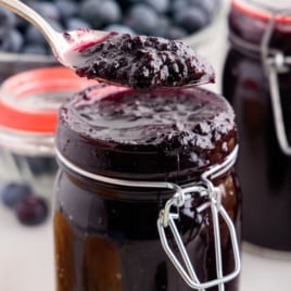Recipe card image featuring blueberry butter on a biscuit with a spoon hovering above, dripping thick, glossy blueberry spread, with glass jars of blueberry butter and fresh blueberries in the background.