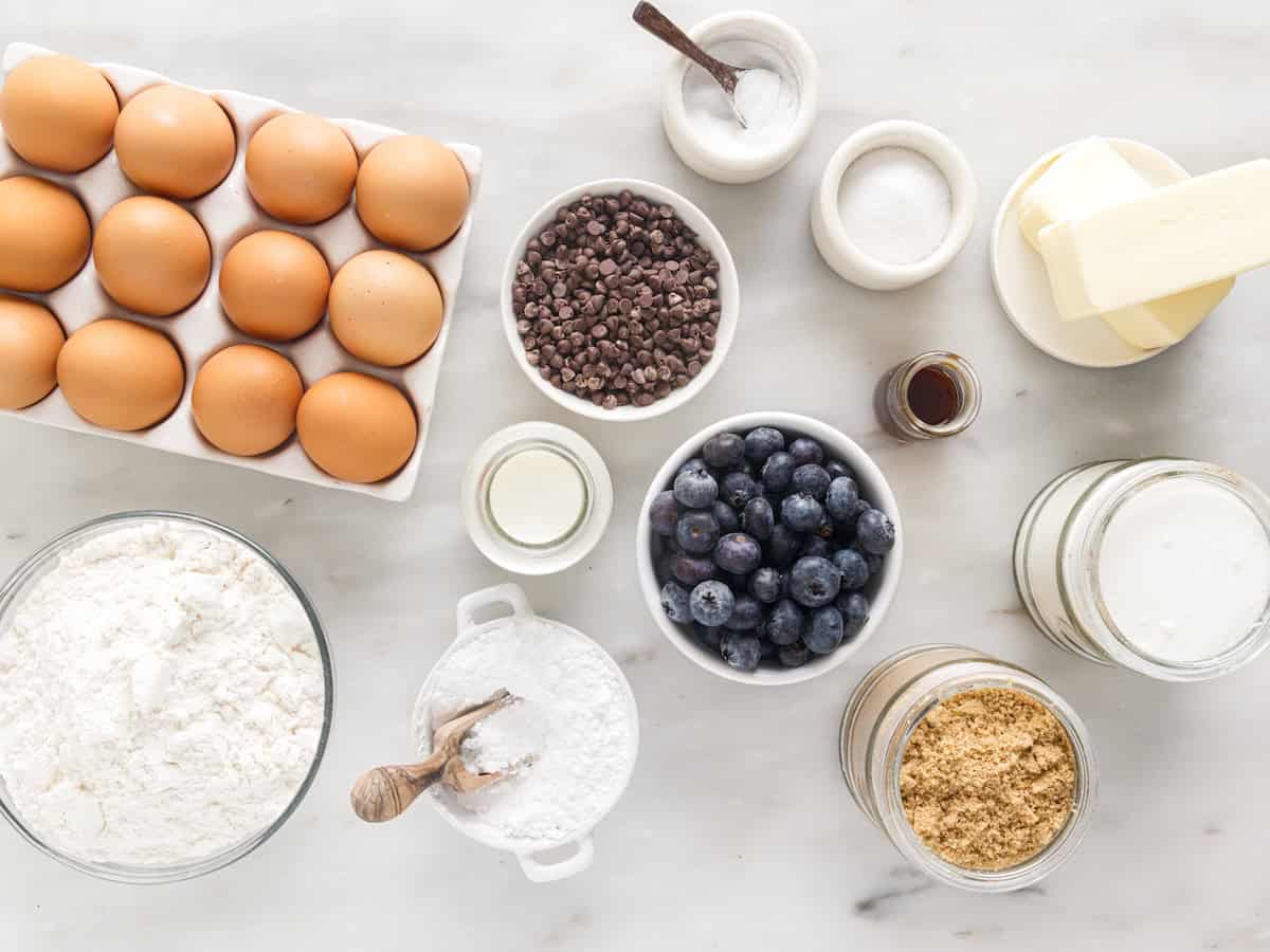 Baking ingredients arranged on a white surface set the stage for delicious Blueberry Chocolate Chip Muffins: eggs, flour, powdered sugar, brown sugar, granulated sugar, blueberries, mini chocolate chips, butter, vanilla, milk, and bowls with salt.