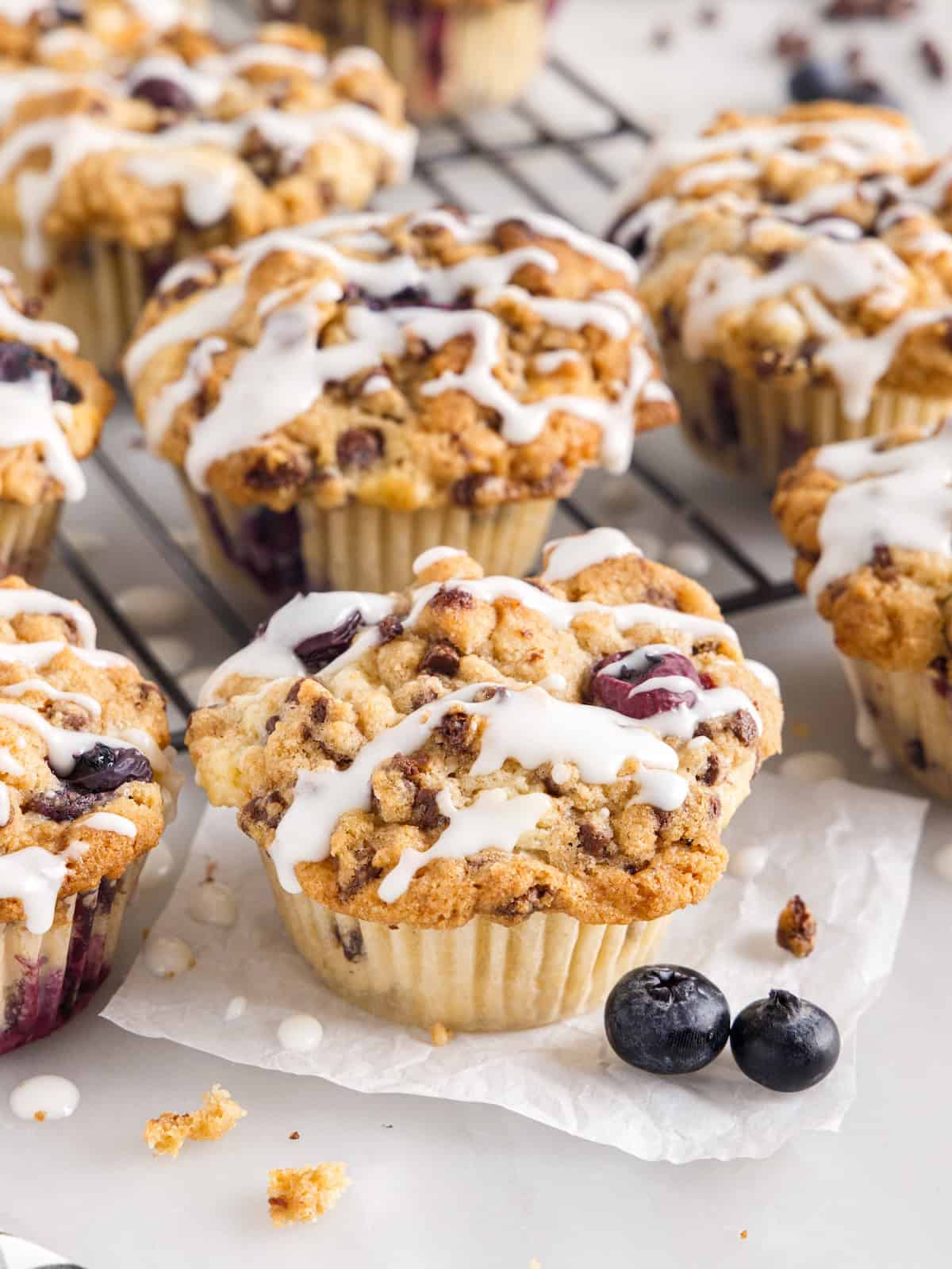 Blueberry Chocolate Chip Muffins with crumb topping and white icing drizzle, arranged on parchment paper and a cooling rack, with fresh blueberries nearby.