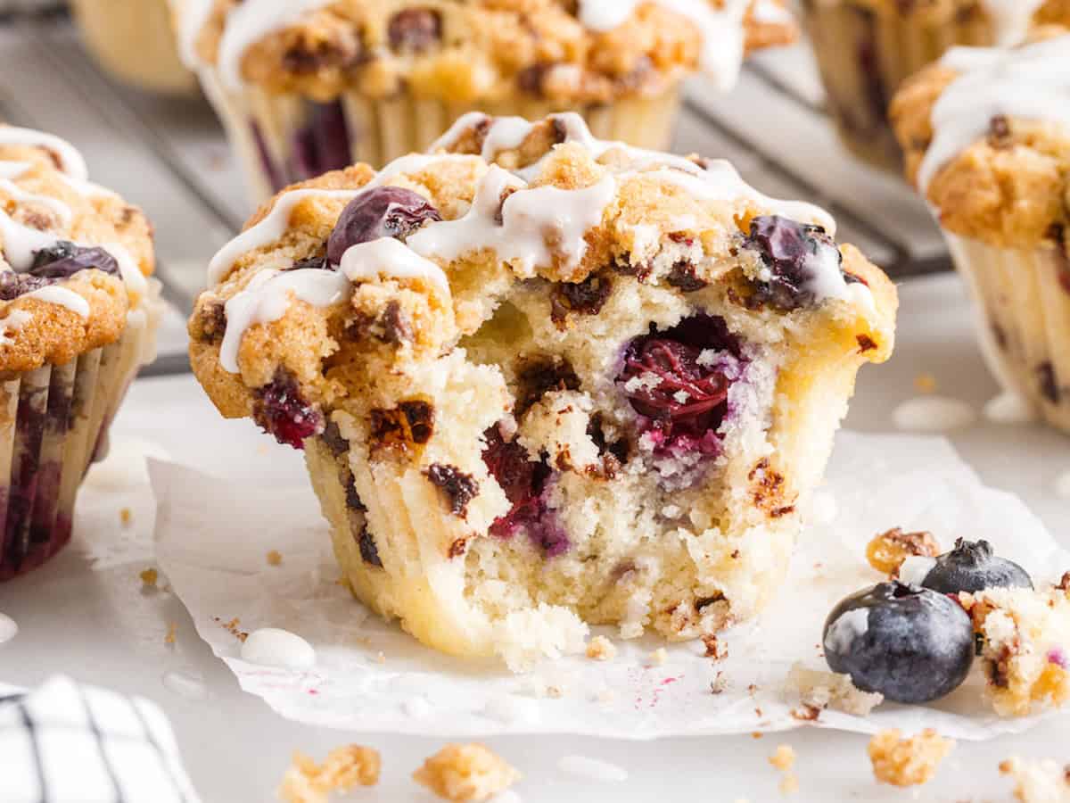 A blueberry muffin with icing sits on parchment paper, broken open to reveal its moist interior and juicy berries. Reminiscent of the classic Blueberry Chocolate Chip Muffins, whole blueberries and muffin crumbs are scattered around.