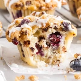 A partially eaten Blueberry Chocolate Chip Muffin with a crumbly topping and white icing sits on parchment paper. Fresh blueberries, chocolate chips, and crumbs are scattered nearby, while other muffins blur softly in the background.
