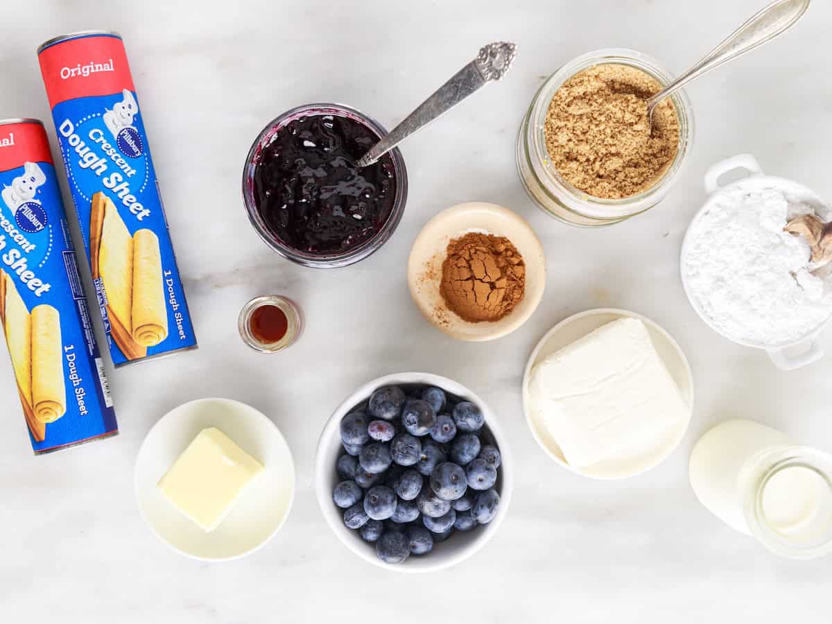 Baking ingredients on a marble surface for Blueberry Cinnamon Rolls: dough sheets, blueberry jam, brown sugar, vanilla, cinnamon, powdered sugar, cream cheese, milk, blueberries, and butter arranged in separate containers.