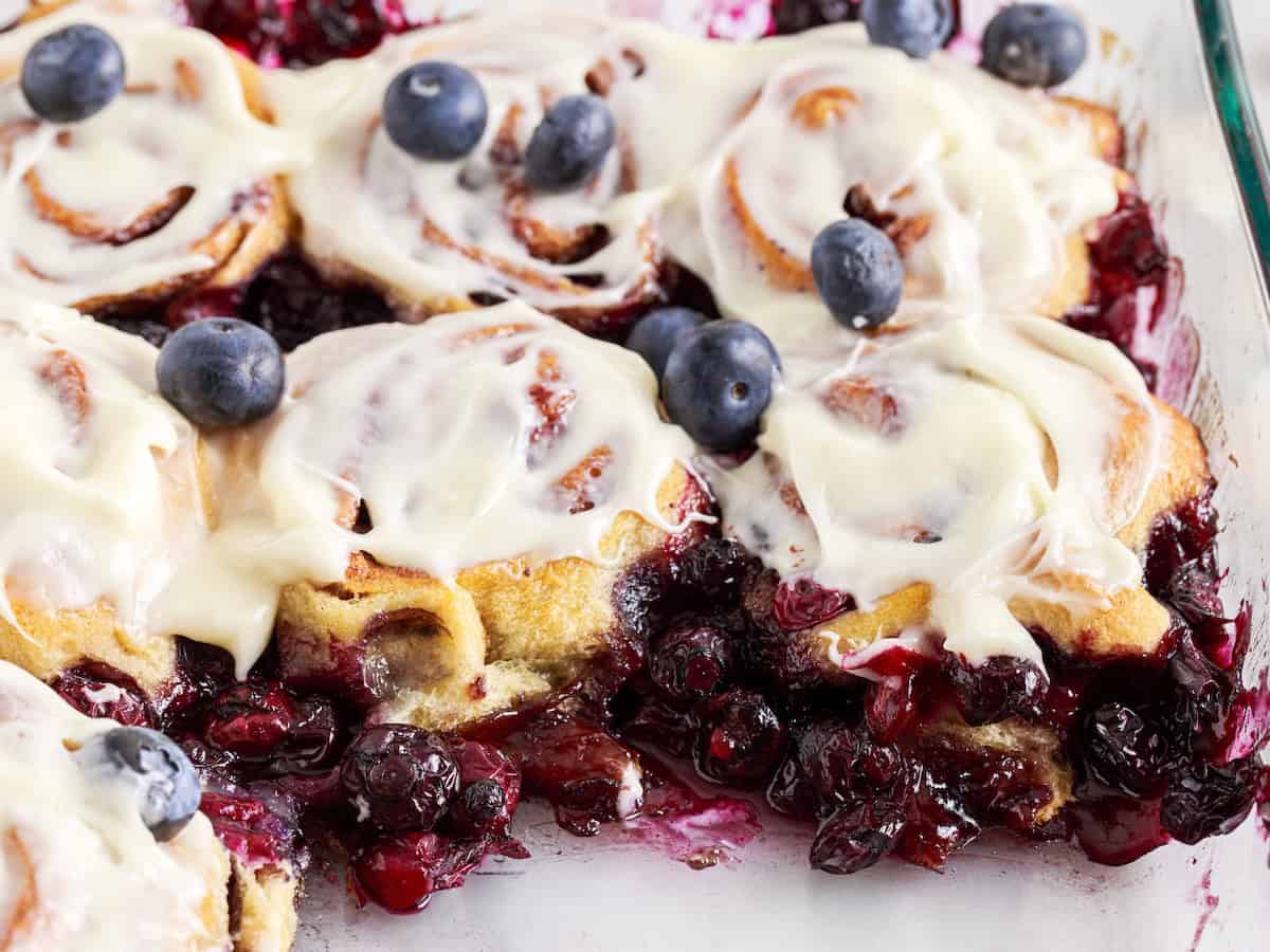 Blueberry Cinnamon Rolls topped with icing and fresh blueberries are baked in a glass dish, with even more berries and luscious berry filling oozing around the warm, fluffy rolls.