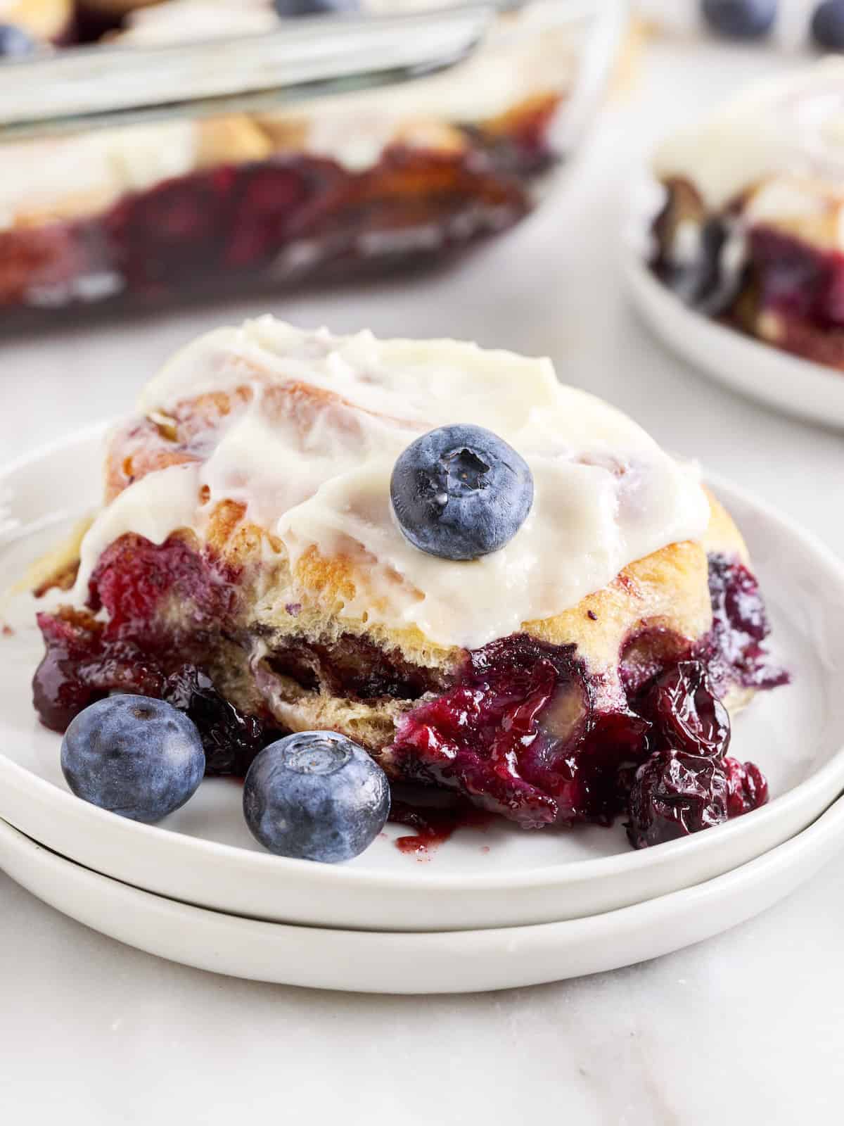 A frosted sweet roll filled with blueberry filling sits on a white plate, topped with a fresh blueberry and surrounded by loose blueberries—an irresistible treat for anyone who loves Blueberry Cinnamon Rolls.