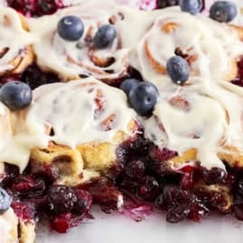 Blueberry Cinnamon Rolls are topped with icing and fresh blueberries, nestled in a glass dish with blueberry filling and sauce oozing around the rolls.