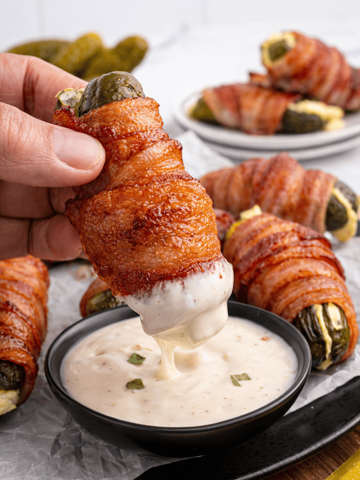 A hand dips a Bacon Wrapped Pickle stuffed with cheese into a bowl of creamy dipping sauce, with more Bacon Wrapped Pickles on a plate in the background.