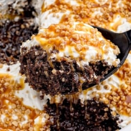 Crockpot Better Than Sex Cake with a spoon lifting a gooey slice of chocolate cake topped with whipped topping, caramel sauce, and crunchy toffee bits inside a white slow cooker.