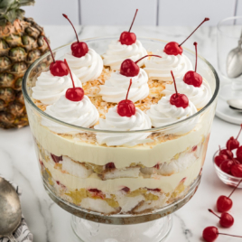 A glass trifle bowl full of layered pina colada trifle. It's garnished with whipped cream and maraschino cherries.