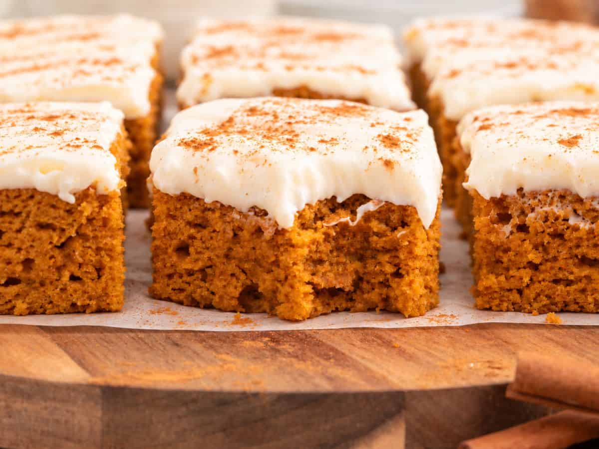 Squares of pumpkin bars topped with white frosting and a dusting of cinnamon on a wooden board.
